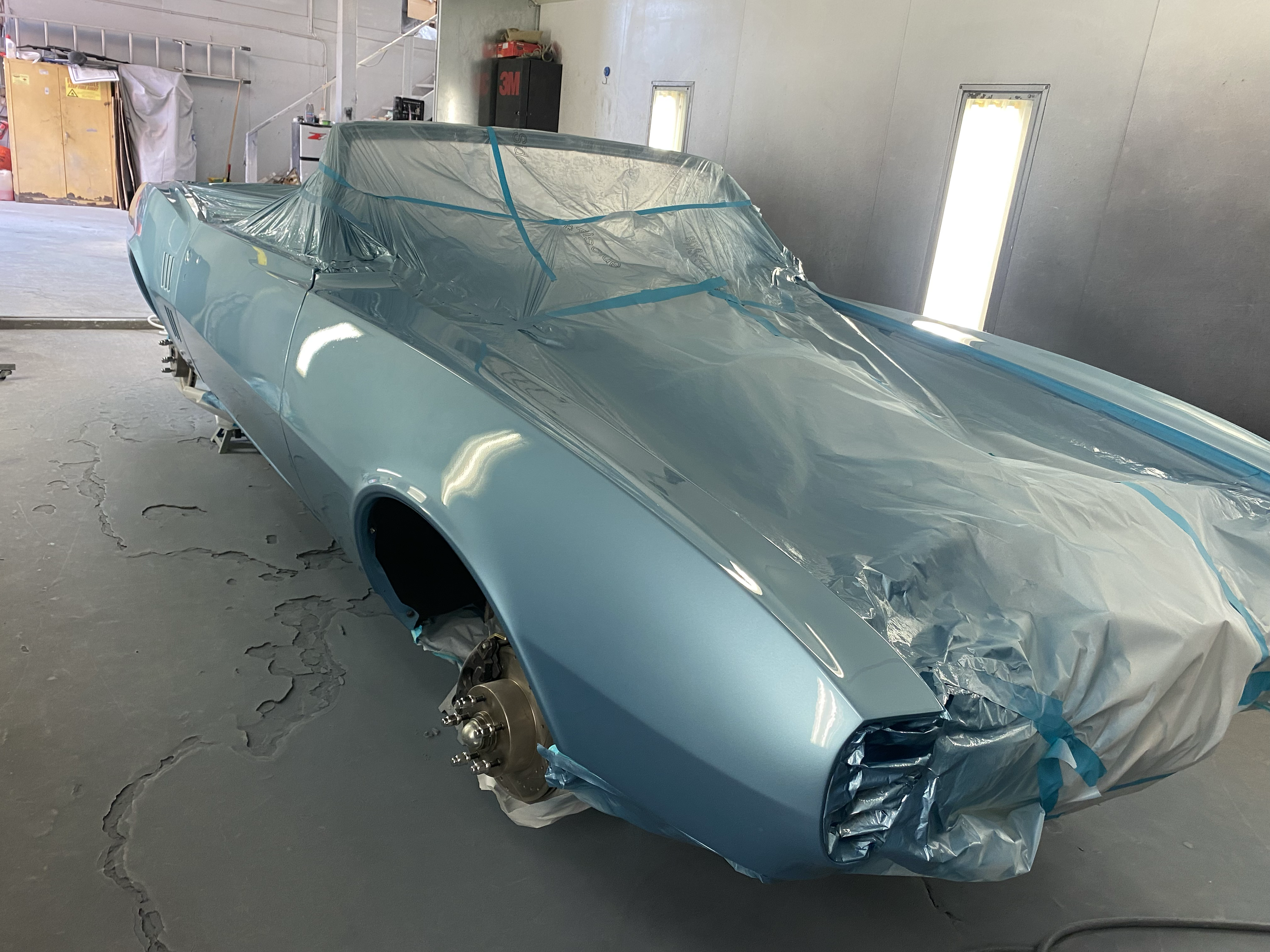 A partially assembled light blue vintage car in a garage, covered with plastic and masking tape, with wheels removed and brakes exposed.