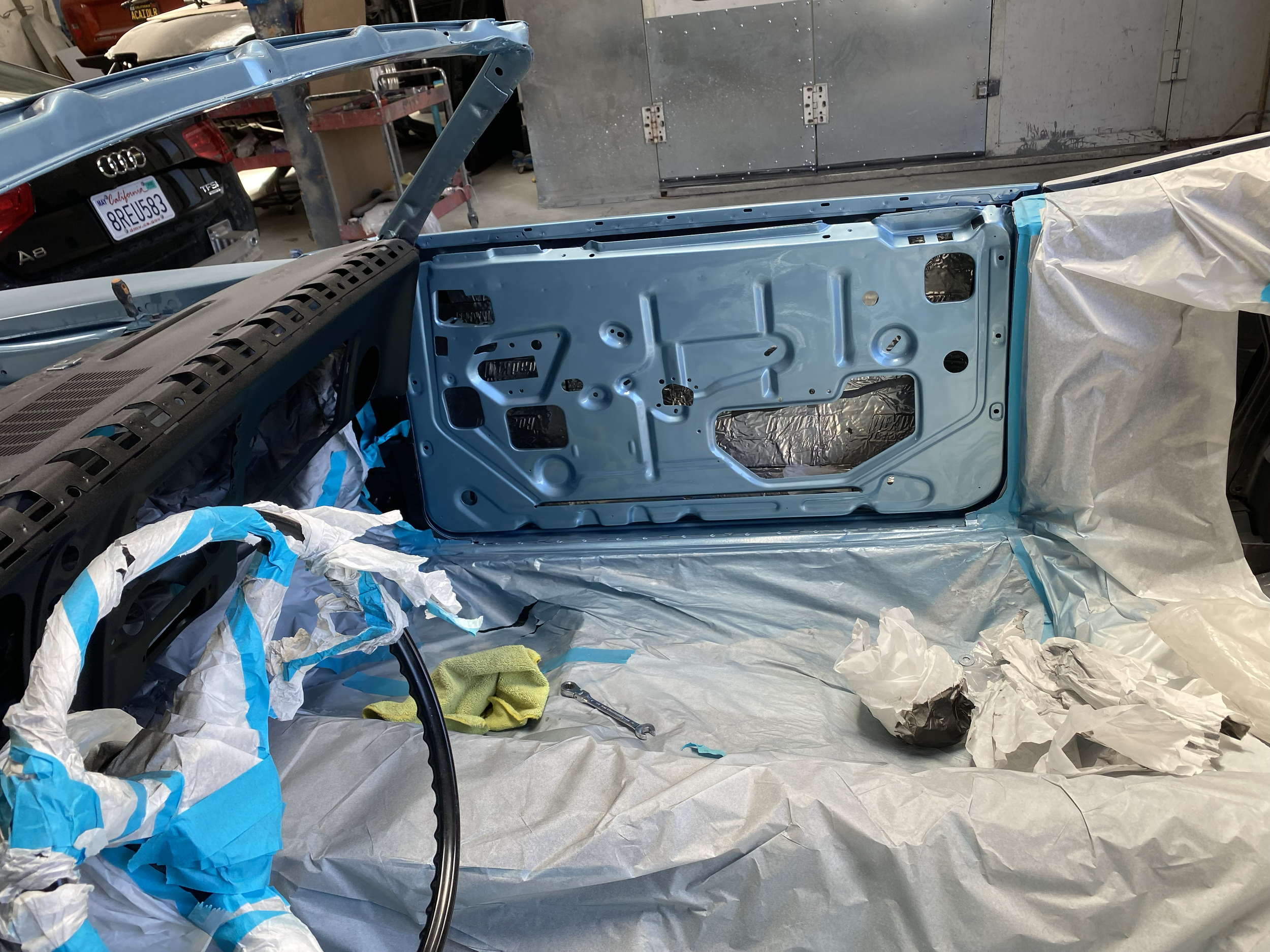 View inside a workshop with a car's car door painted in light blue. The door is removed from the vehicle and laid flat on a surface. Surrounding the door are crumpled paper and masking tape used for painting. In the background, a black car with Calif