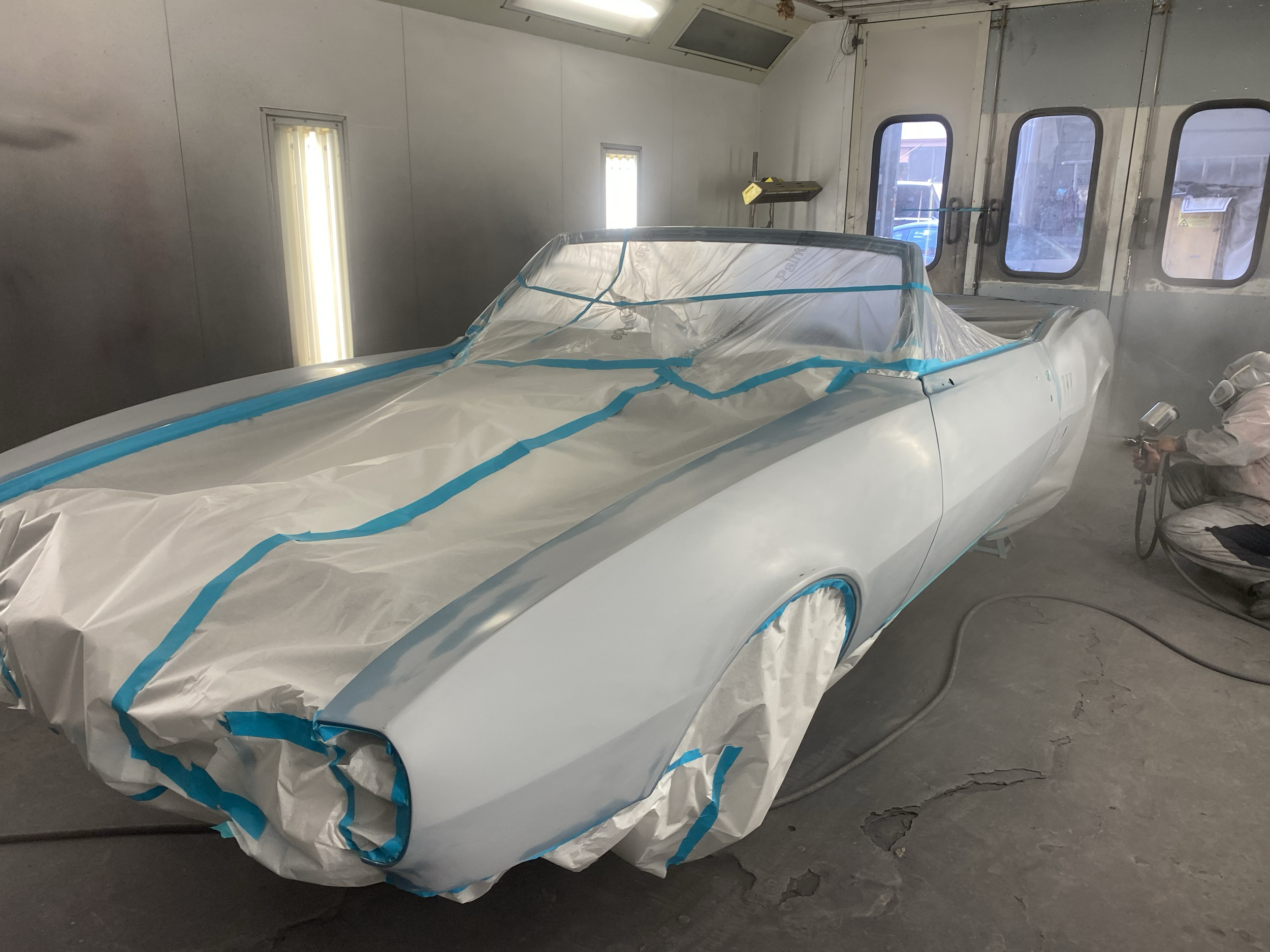 A vintage white sports car covered with protective paper and blue painter's tape inside an industrial, unpainted garage or workshop with windows and a door.
