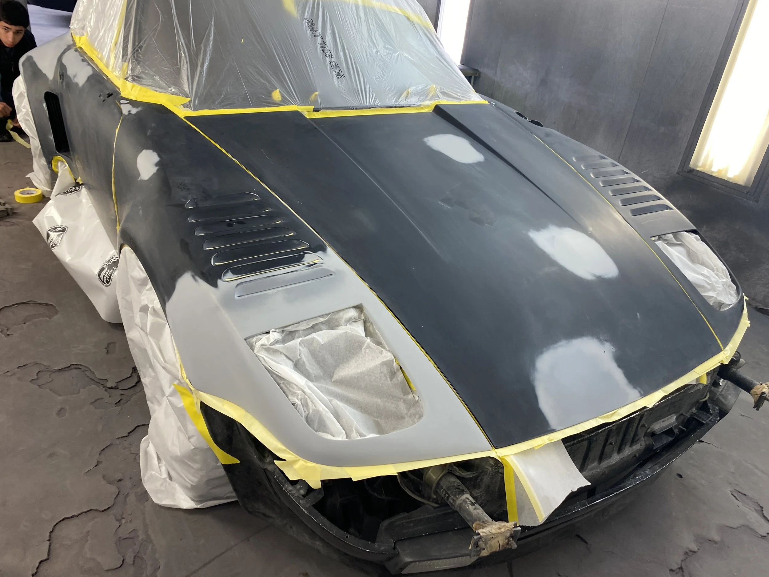 A car being prepared for painting or finishing, covered with masking tape and paper, with parts of the bodywork in black and unpainted areas in primer or bare metal, inside a workshop.