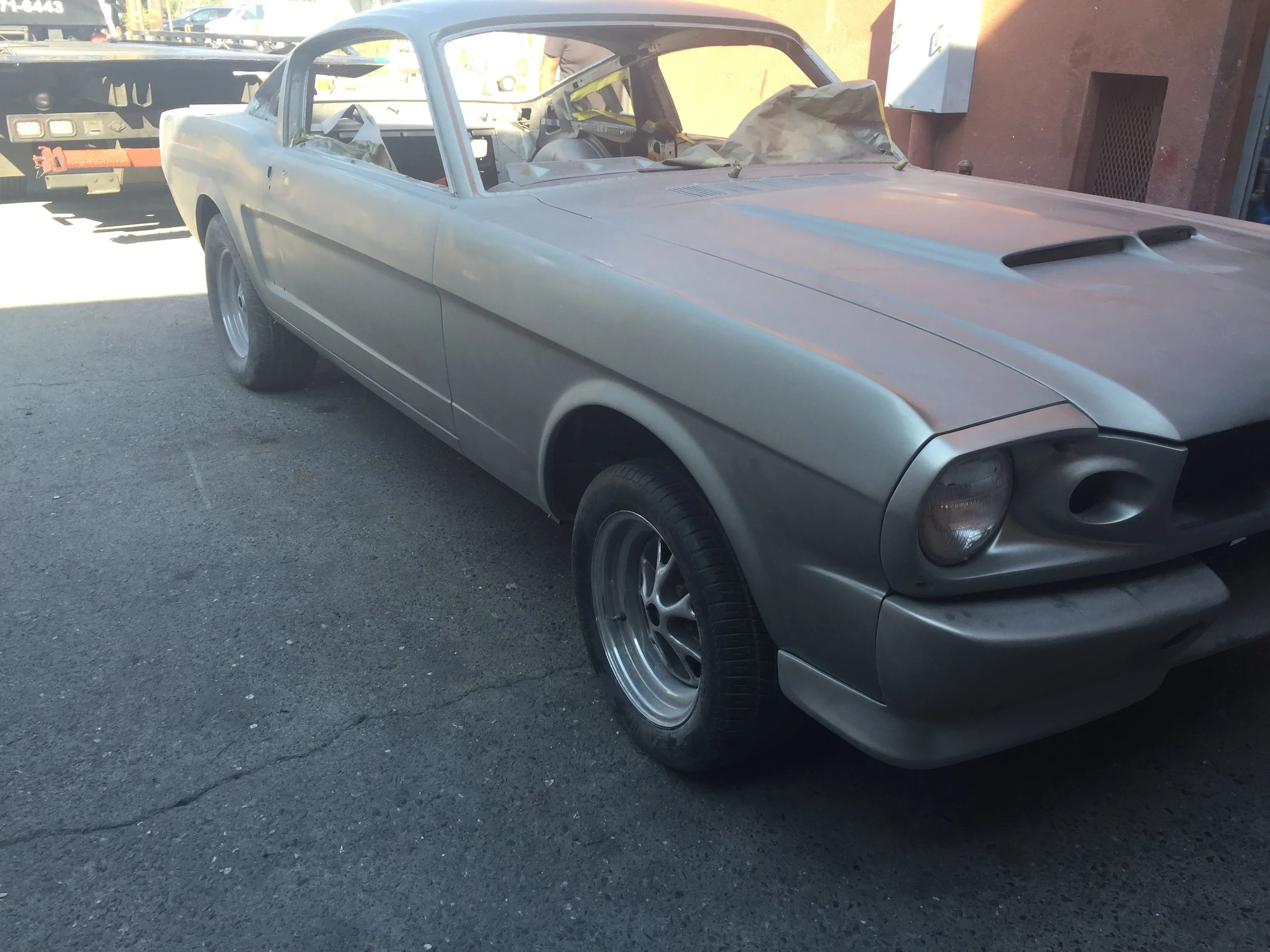 An unfinished or vintage silver or gray car with a sleek design, missing interior parts, parked on a paved surface next to a red building, with some objects and vehicles in the background.