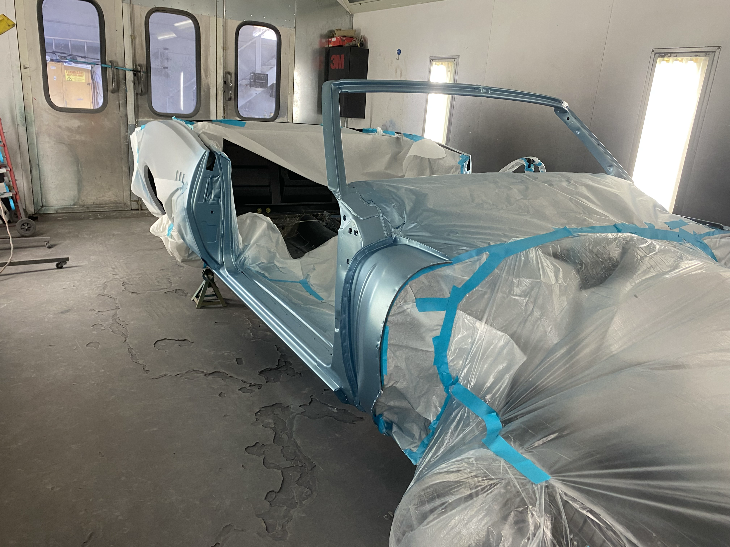 A partially assembled car body in a spray booth, covered with plastic or masking paper and blue painter's tape, with a metal tool cart and equipment in the background.