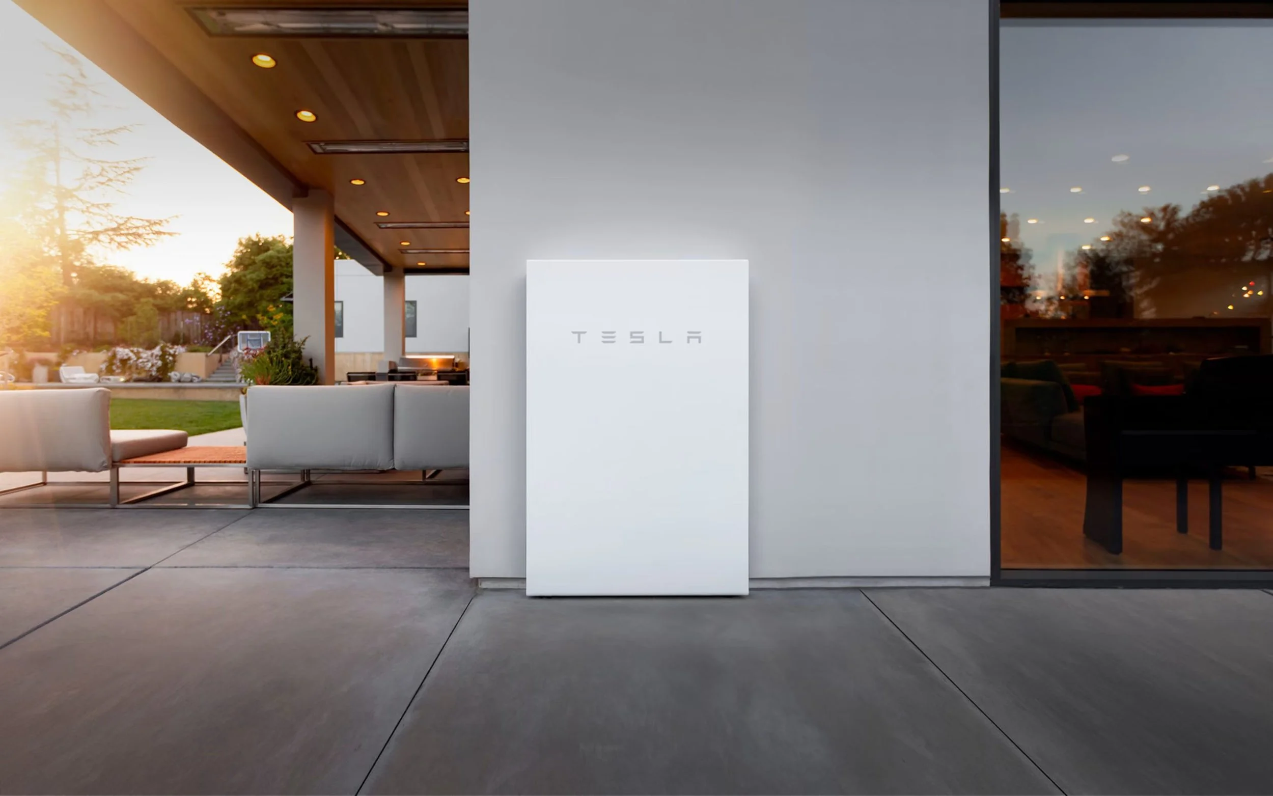 Image showing the exterior view of a Tesla showroom at sunset, with a white Tesla display stand in the center, large glass windows revealing a modern interior, outdoor seating, and greenery in the background.