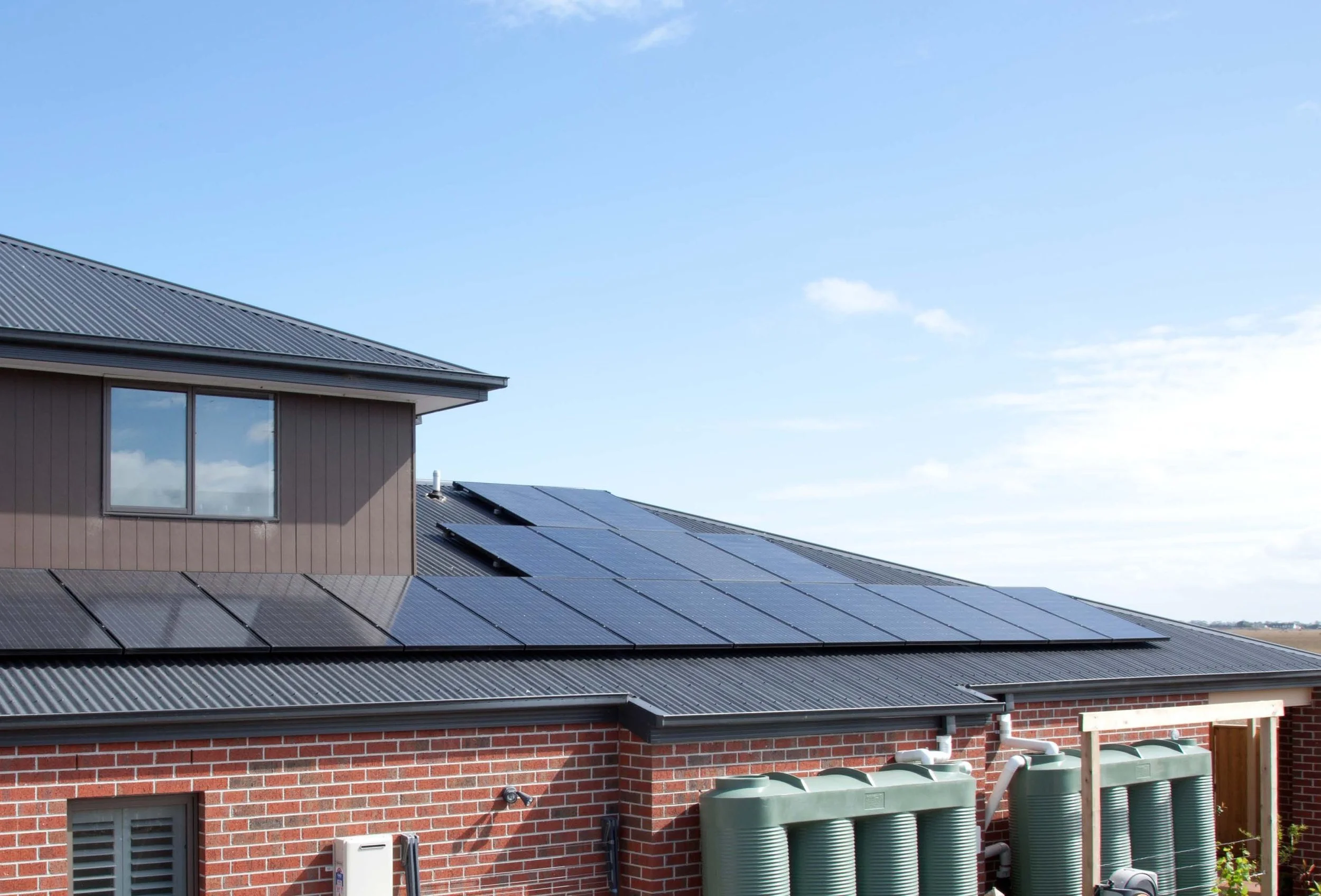 A house with solar panels on the roof, a window, a brick wall, and several outdoor utility boxes and water tanks under a blue sky with some clouds.