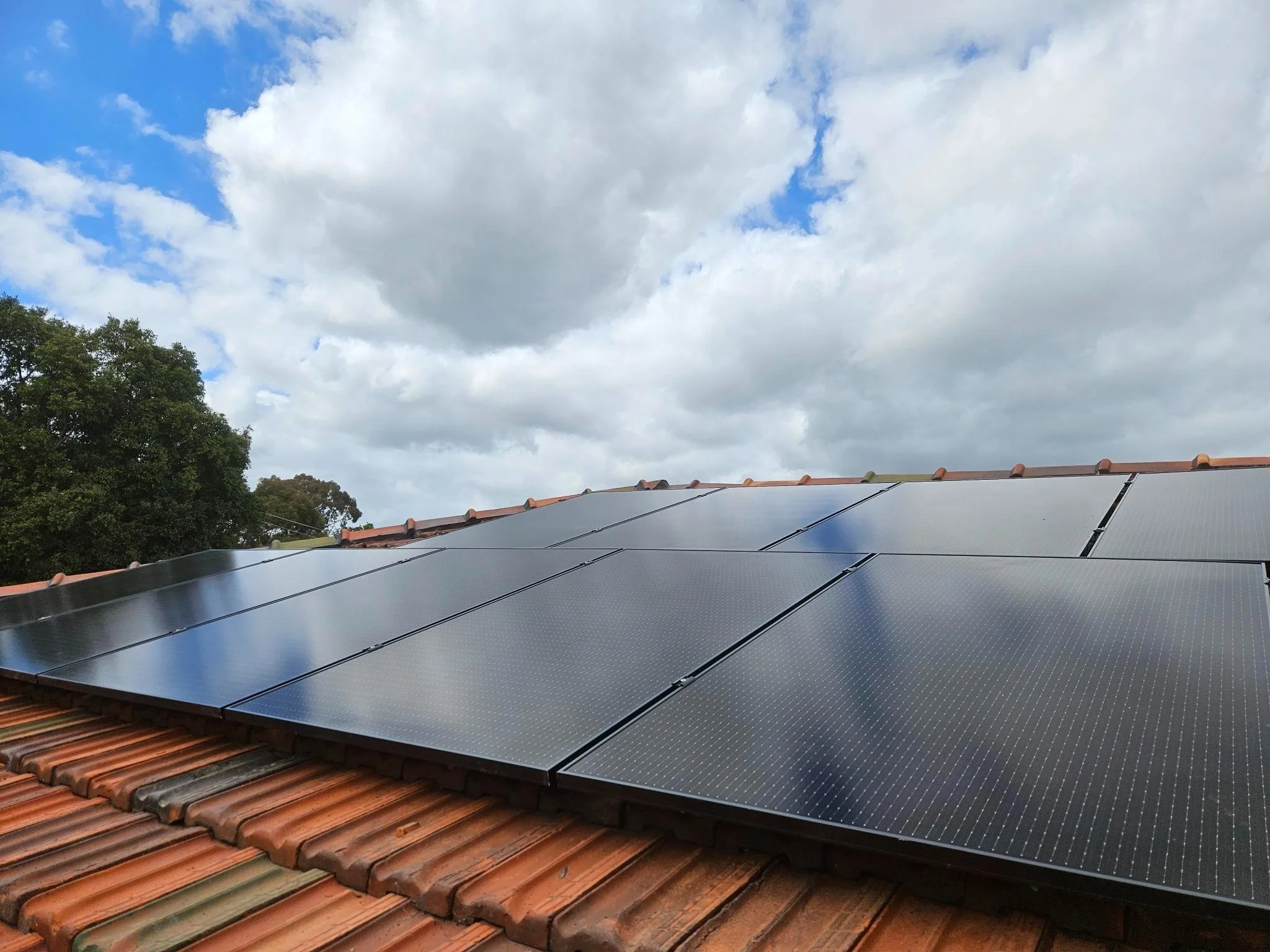 Solar panels installed on a house roof with terracotta tiles, under a partly cloudy sky with some green trees in the background.