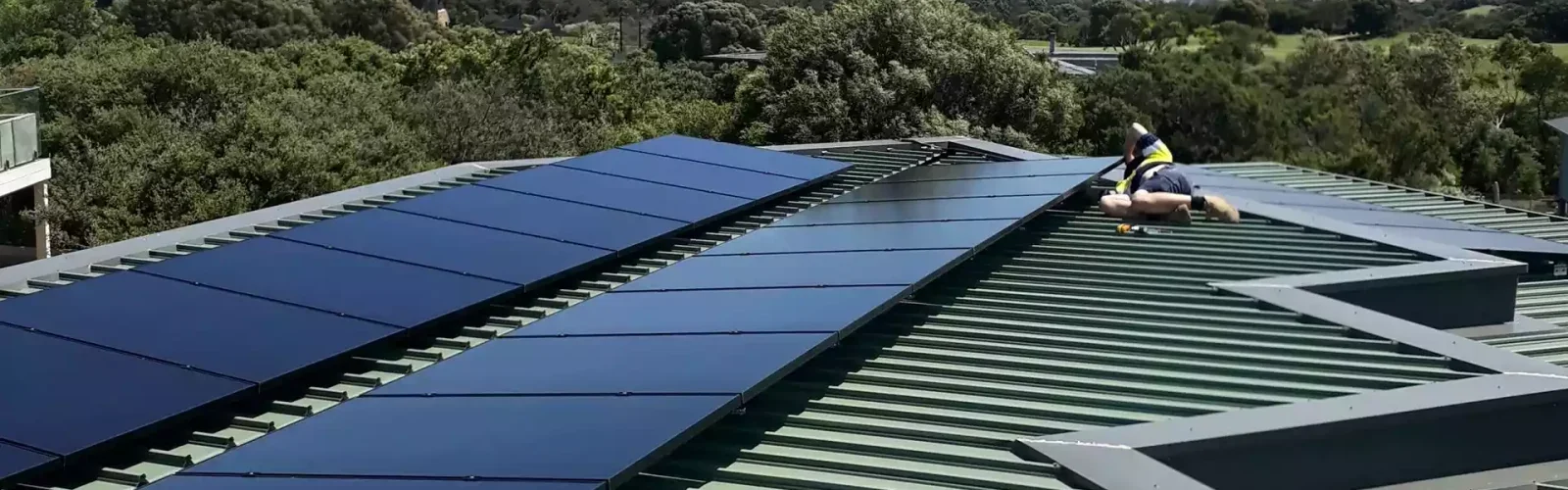 A worker installing solar panels on a metal roof in a residential area, surrounded by trees and other houses.