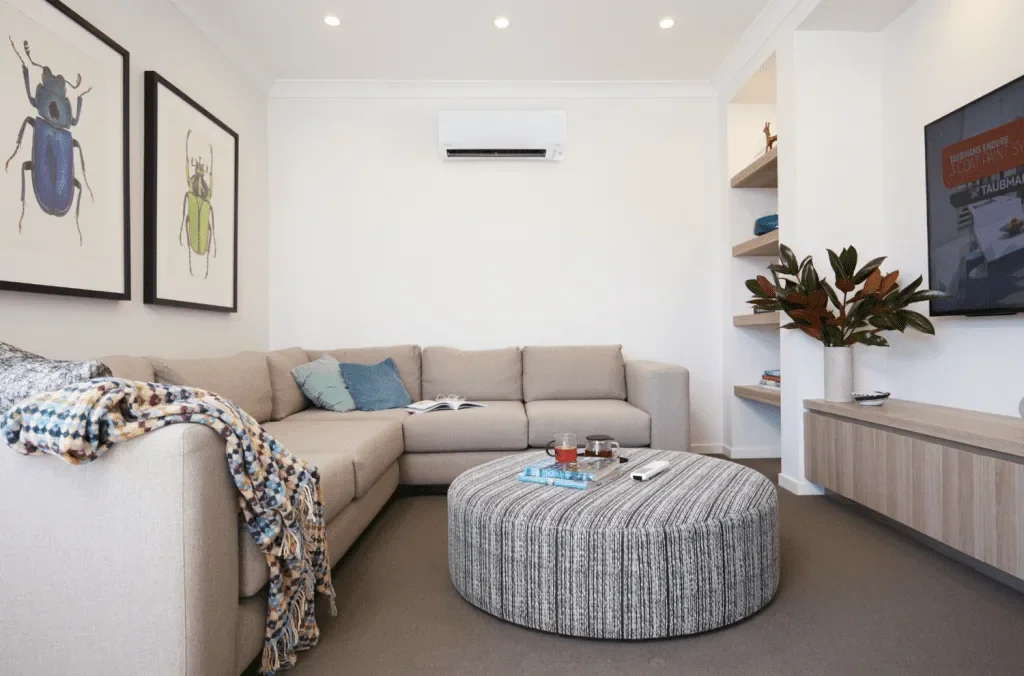 Living room with a beige L-shaped sofa, colorful throw blanket, blue and gray pillows, a round striped ottoman with drinks, a wall-mounted TV, framed insect art, built-in shelves with decor, and a large plant in a white vase.