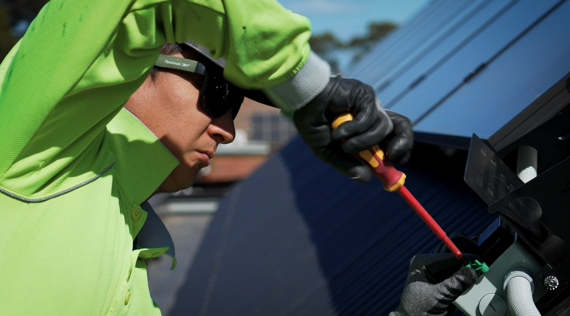 A man wearing a neon yellow jacket, black gloves, and sunglasses is working on a solar panel with a screwdriver.
