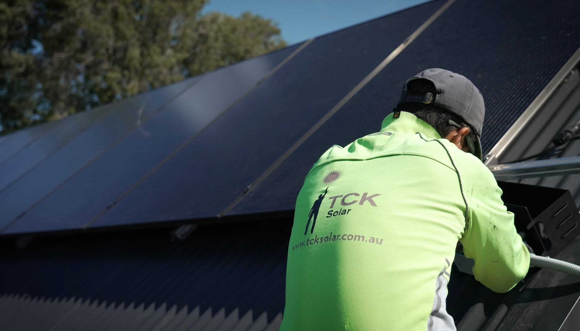 A worker in a bright yellow long-sleeve shirt and gray cap installing solar panels outdoors on a sunny day, with trees and a blue sky in the background.