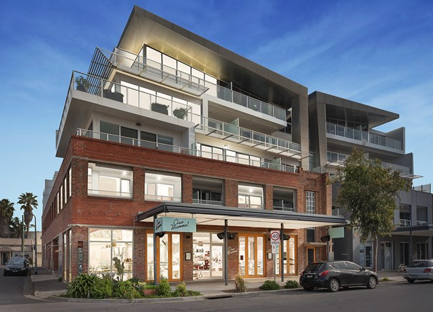 Modern multi-story building with retail space on the ground floor and residential units above, featuring brick and glass design and a small landscaped area at the entrance, under a clear blue sky.