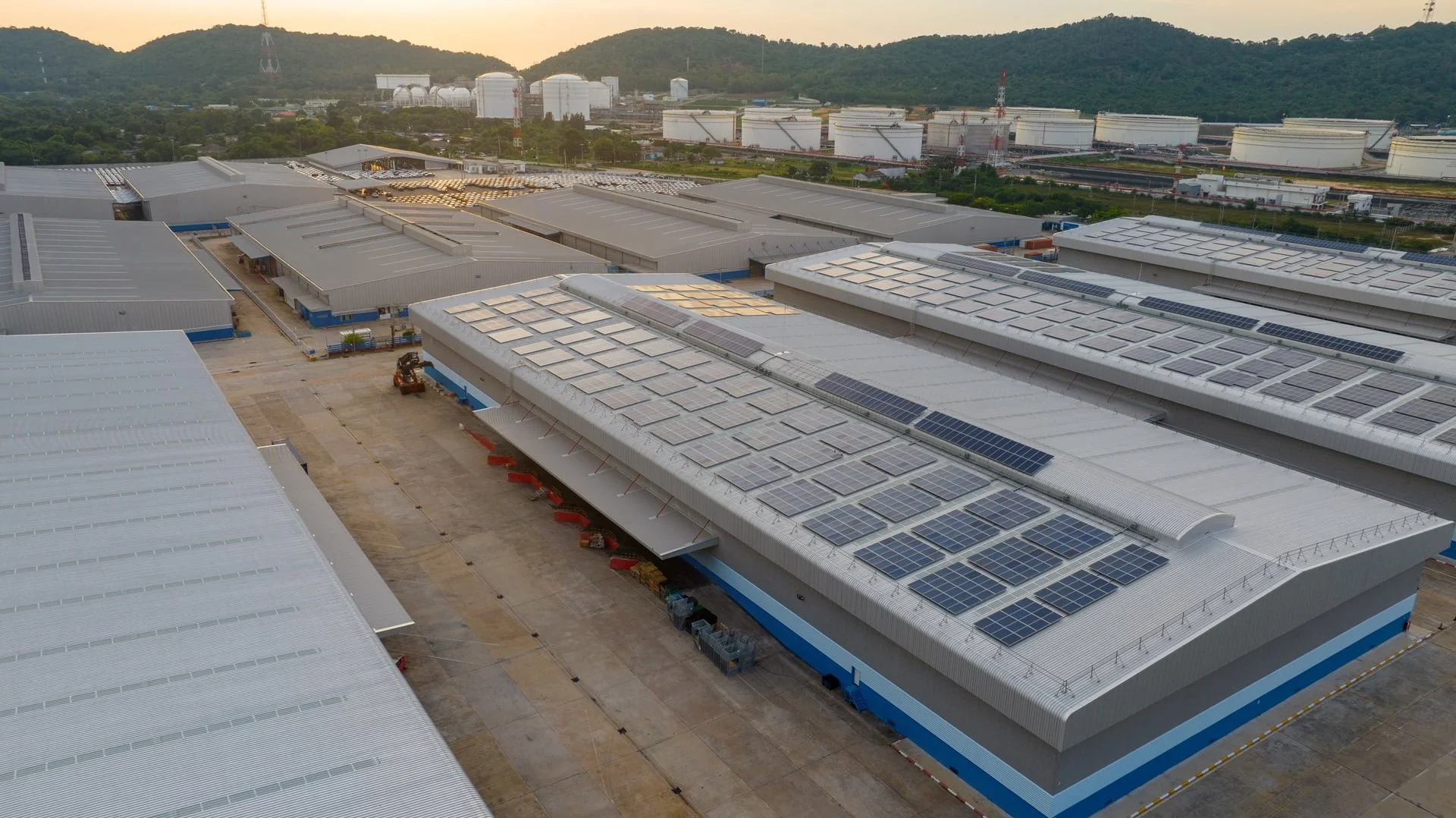 Aerial view of a large industrial warehouse complex with solar panels on the roofs, surrounded by mountains in the background at sunset.