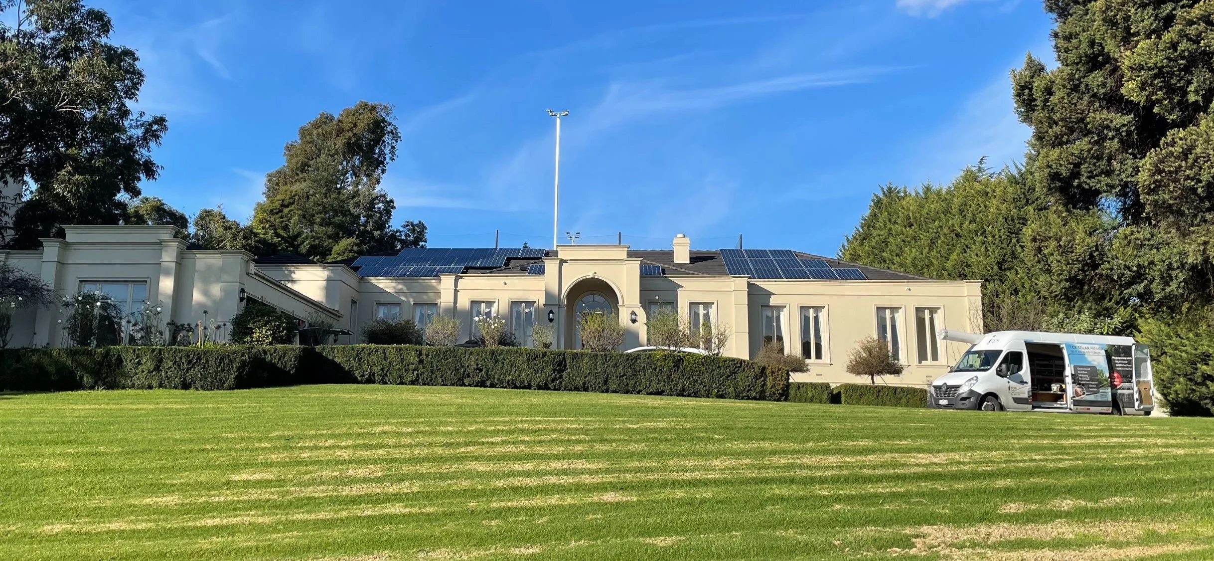 A large beige house with solar panels on the roof, surrounded by green trees and bushes, with a well-manicured lawn in the foreground. A white service van is parked near the house.