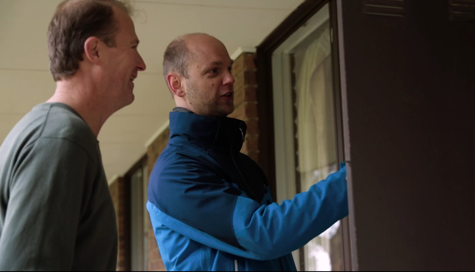 Two men are standing outside a house near a window. One man is wearing a gray shirt and smiling, while the other man, dressed in a blue jacket, is reaching out to a bulletin board or noticeboard.