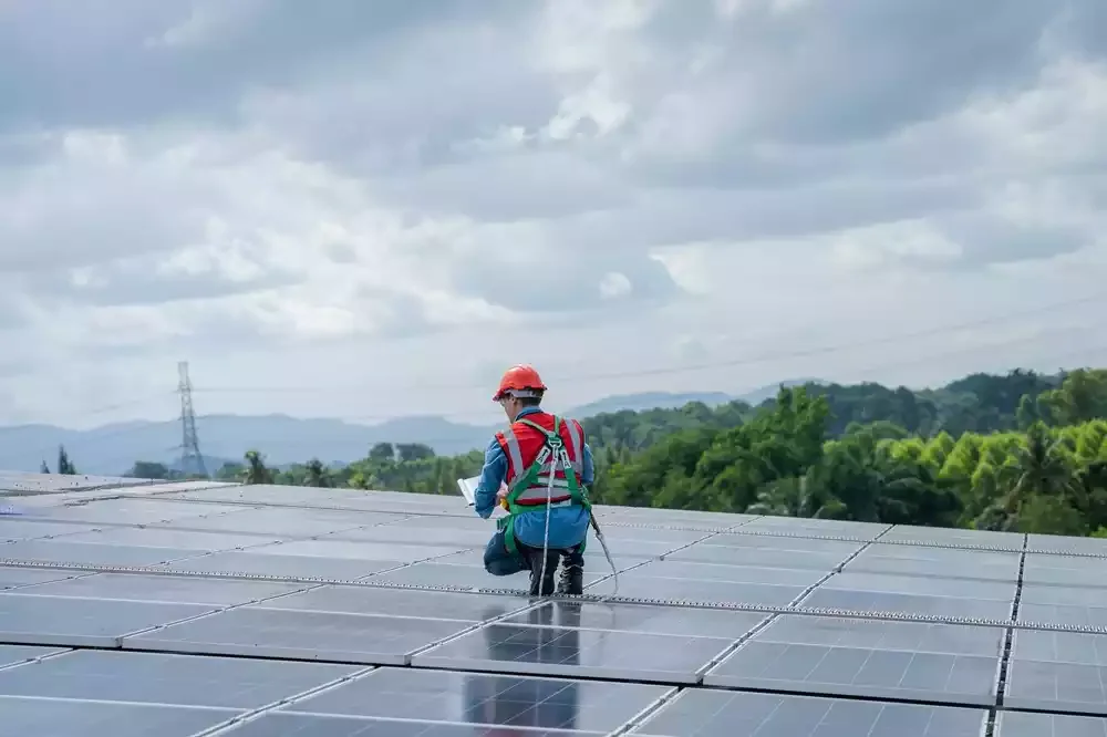A person wearing a red safety helmet, harness, and backpack kneeling on solar panels in an open solar farm with greenery and cloudy sky in the background.