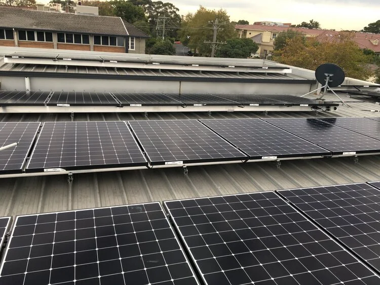Multiple solar panels installed on a building roof with houses and trees in the background.