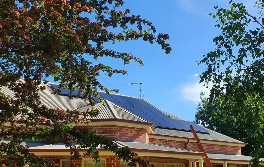A house with a brick exterior and a metal roof, featuring solar panels, surrounded by trees with green and reddish leaves, under a blue sky.