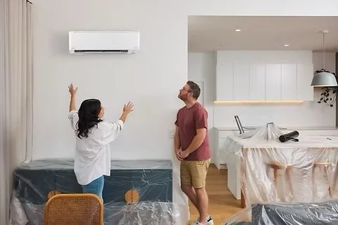 Two people standing in a modern living room, talking and gesturing at each other, with an air conditioning unit on the wall.