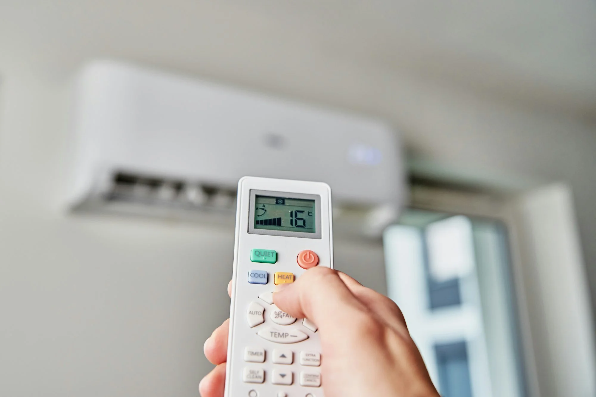 Person holding a remote control in front of a wall-mounted air conditioner with a digital display showing the temperature set to 16°C.