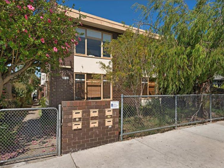 Two-story residential building with a fenced front yard, mailboxes, trees, and a sidewalk.