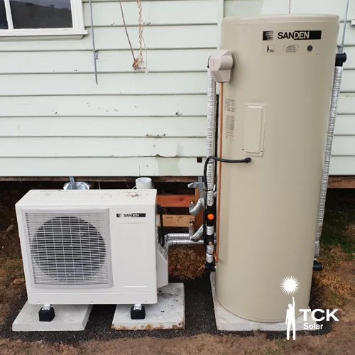 An outdoor sanden heating or cooling system installation, with a large vertical storage tank and an air conditioning or heat pump unit, connected by pipes, in front of a house with horizontal siding.