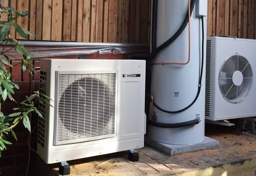 Exterior of an air conditioning system with two outdoor units and a heat pump, installed next to a brick wall and wooden fence, with some greenery in the foreground.