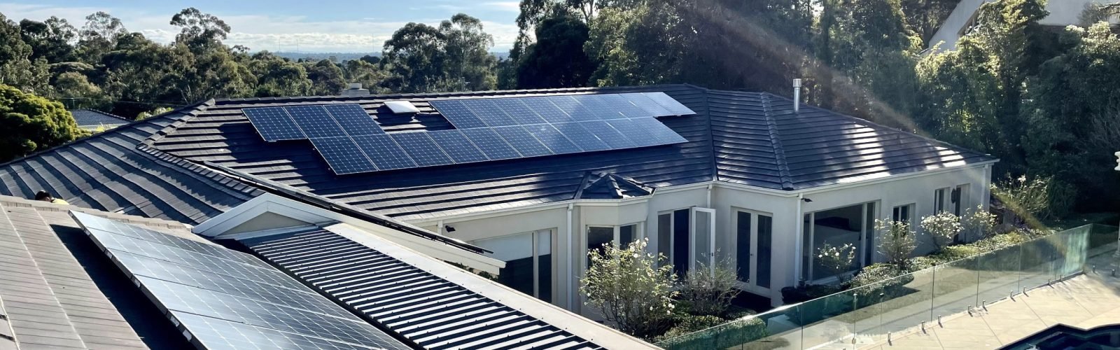 Residential house with solar panels on the roof, surrounded by trees and greenery, with a clear sky and sunlight.