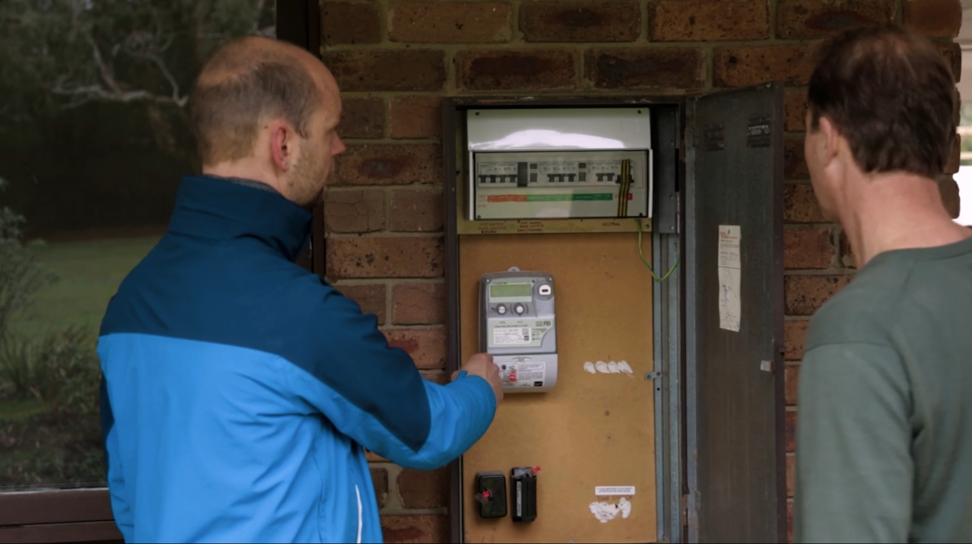 Two men are working on an outdoor electrical meter and breaker box mounted on a brick wall. One man is using a handheld testing device near the meter. The other man is observing. The scene is outside next to a wall with some greenery visible.