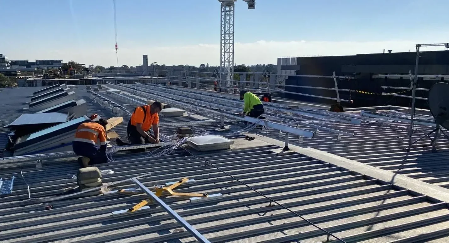 Construction workers installing solar panels on a building roof, with rooftop equipment and a crane in the background.