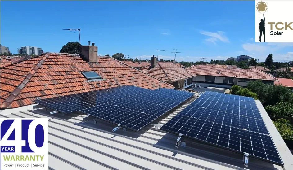 Solar panels installed on metal roof of a residential building with red tiled roofs and a clear blue sky in the background.