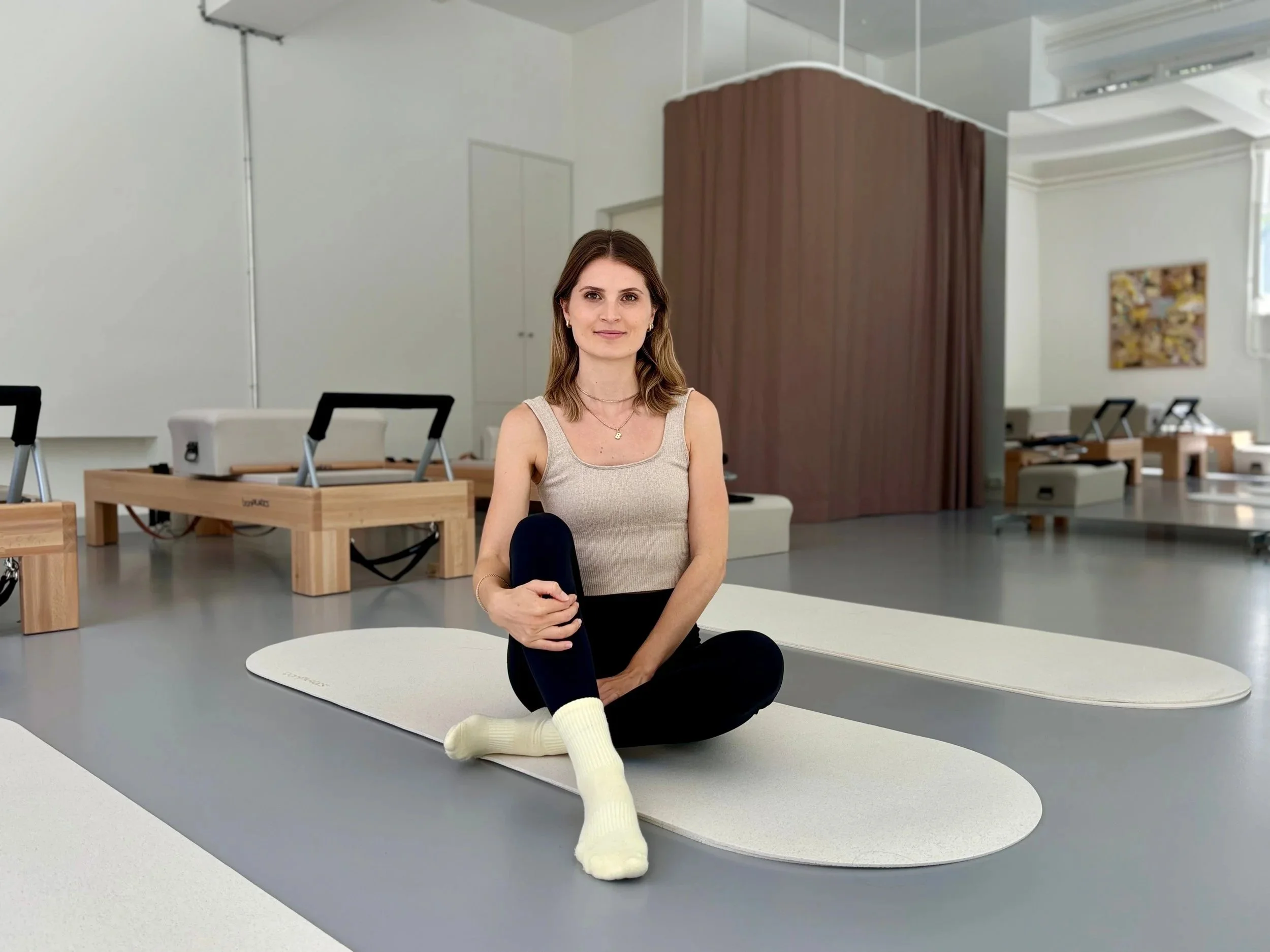 Junge Frau mit dunkelblonden mittellangen Haaren sitzt in beige-schwarzen Sportbekleidung auf einer Pilates-Matte in einem Pilatesstudio. Im Studio sind schöne braune Vorhänge und Pilates-Geräte, Reformer, sowie Rollspiegel m Hintergrund zu sehen.