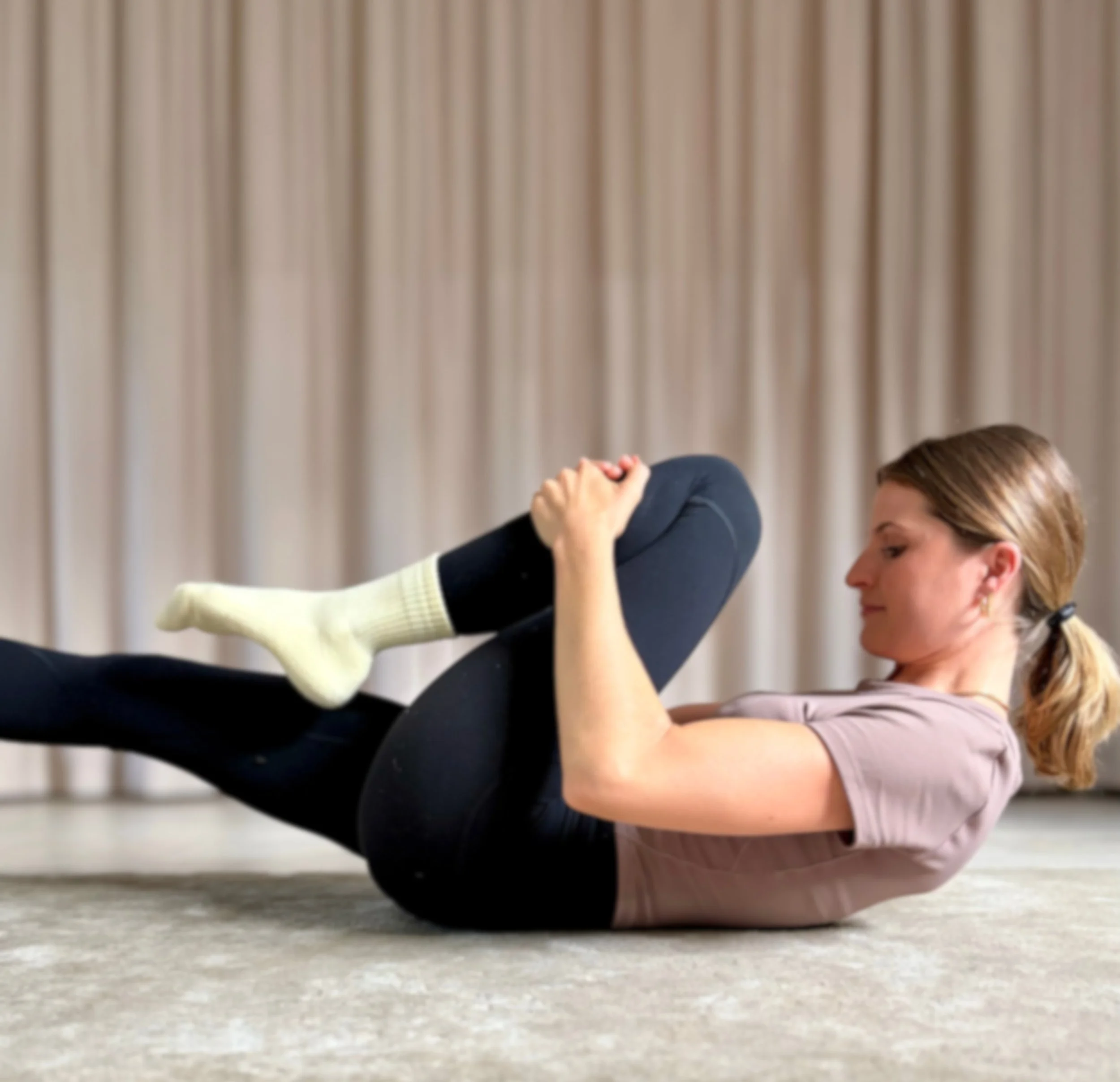 Frau macht Pilates-Übung auf der Matte in einem Raum mit Vorhängen im Hintergrund.