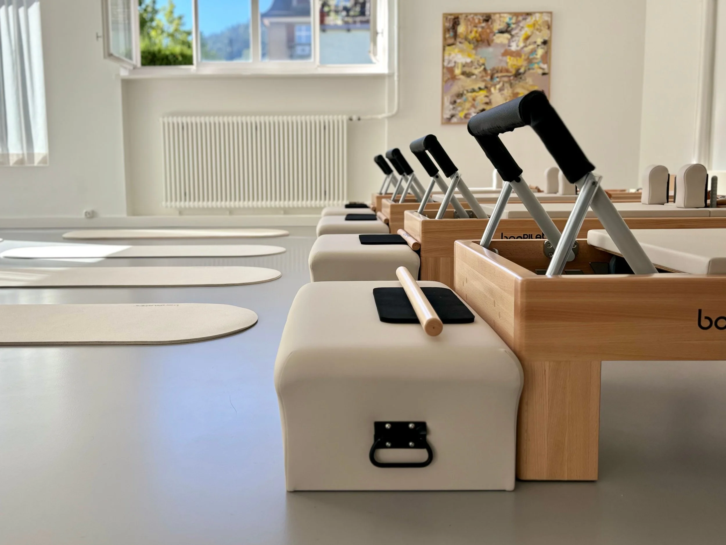 Vier Pilates-Geräte, Reformer, stehe in einer Reihe vor einem hellen grossen Fester und einem bunten Bild, das an der Wand befestigt ist. Vor den Reformern liegen Pilates-Matten auf dem Boden.