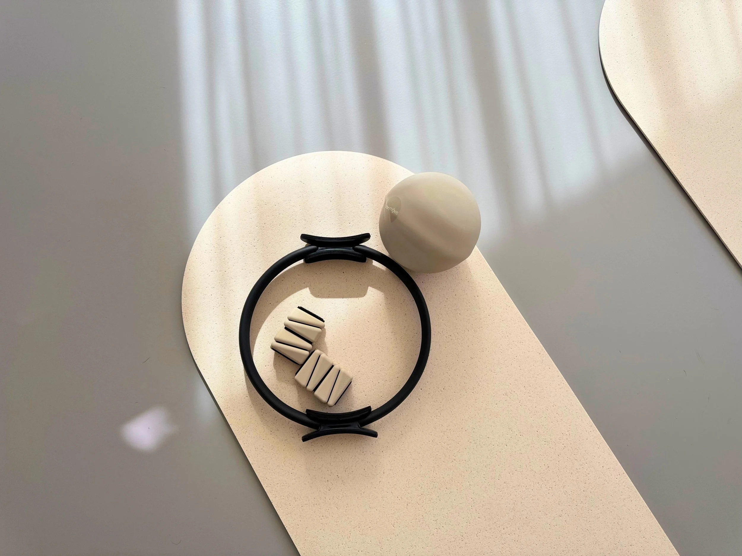 Pilates-Equipment: Beige Matte, grauer Pilates-Ball, schwarzer Pilates-Ring und beige Gewichte für die Fussgelenke. Die Sonne strahlt auf das Equipment.