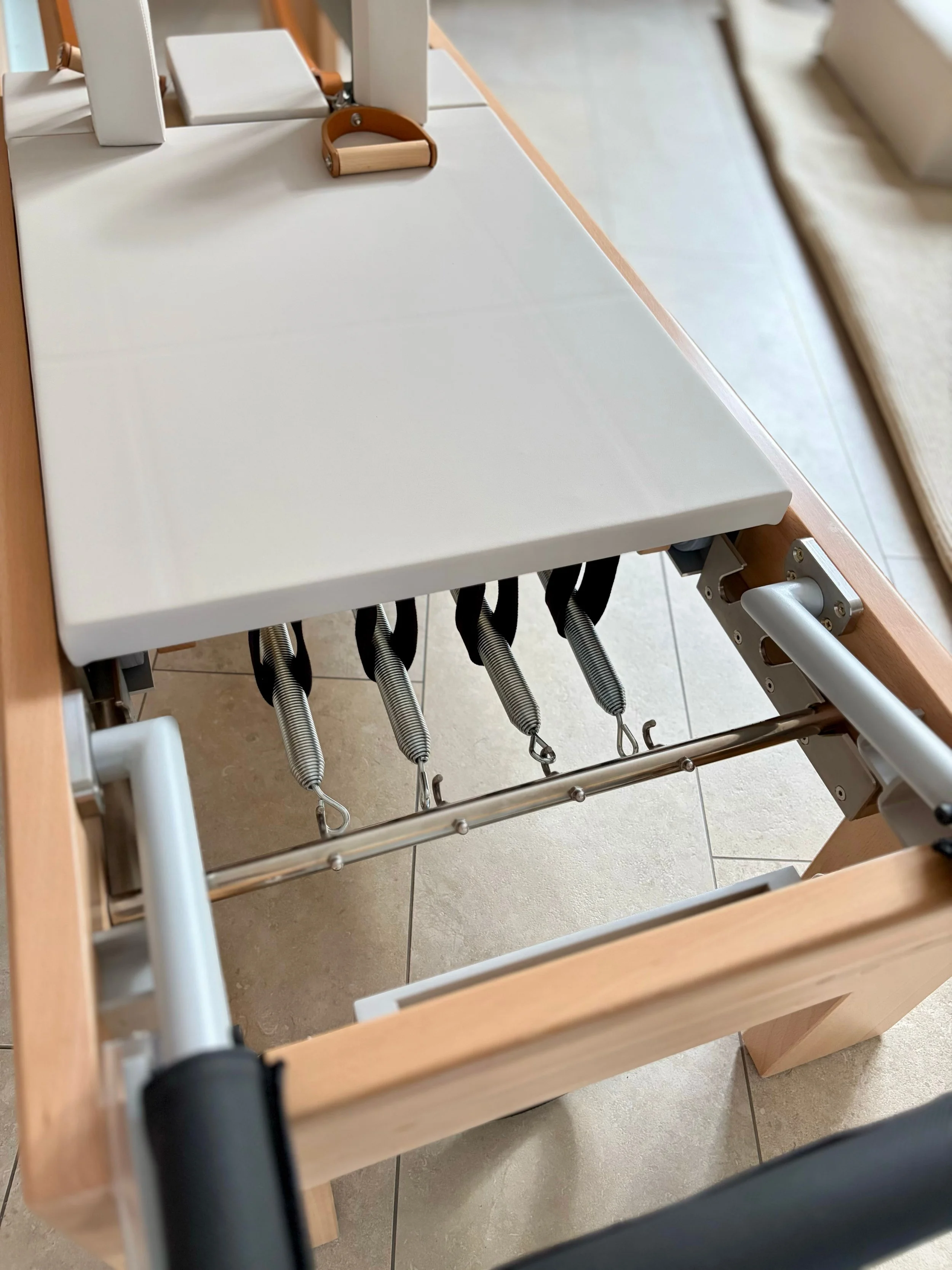 Ein Pilates Gerät mit weissen Polstern, braunen Ledergriffen, Metallstützen mit Holzrahmen.