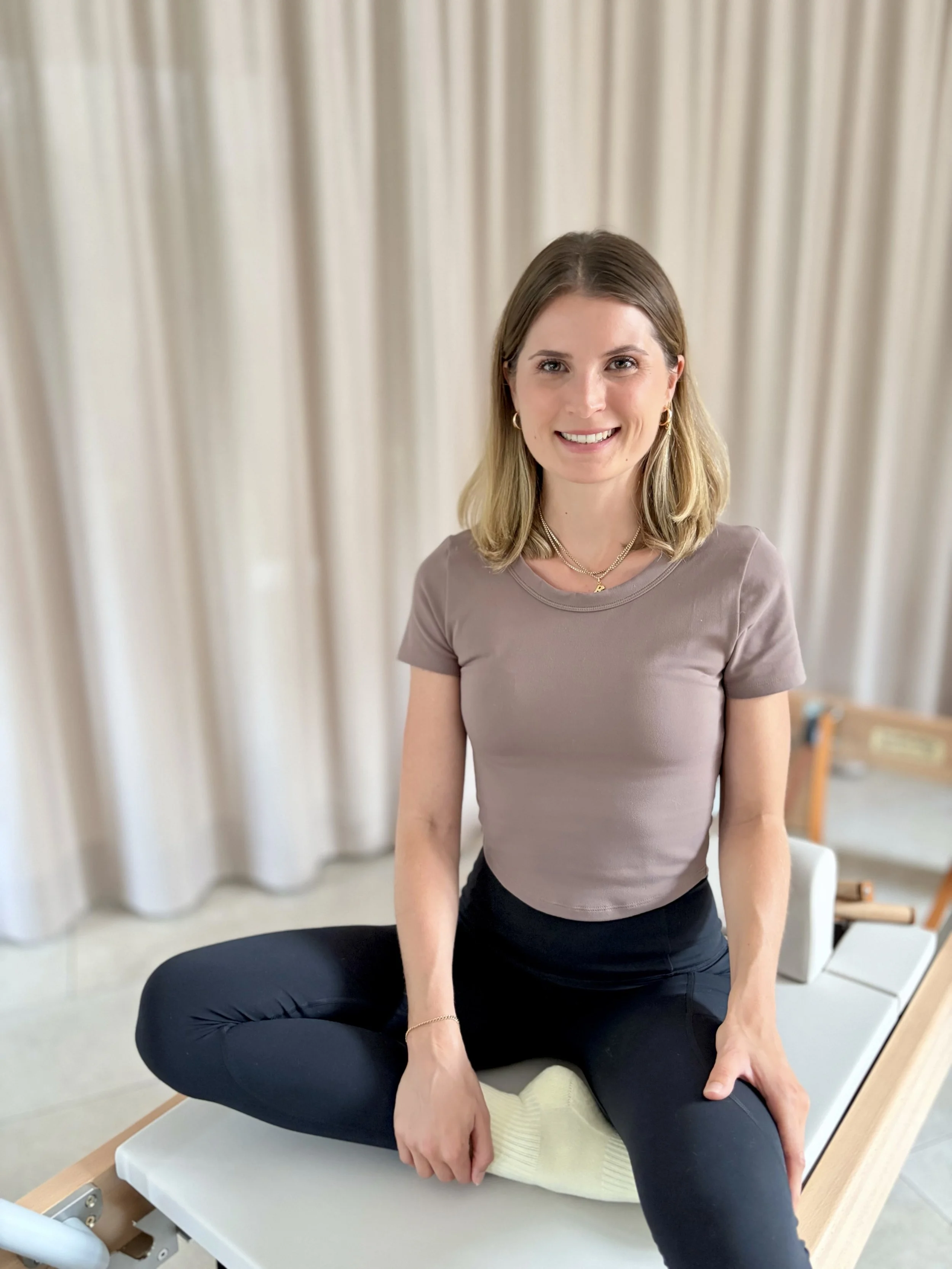 Frau in sportlicher Kleidung sitzt auf einem Pilates Reformer in einem Raum mit weißen Vorhängen im Hintergrund.