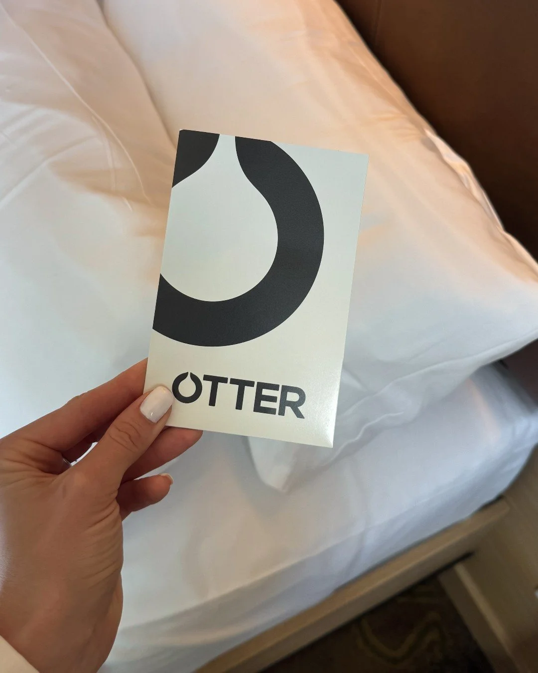 Hand holding an OTTER card with a bold black abstract logo over a white bed.
