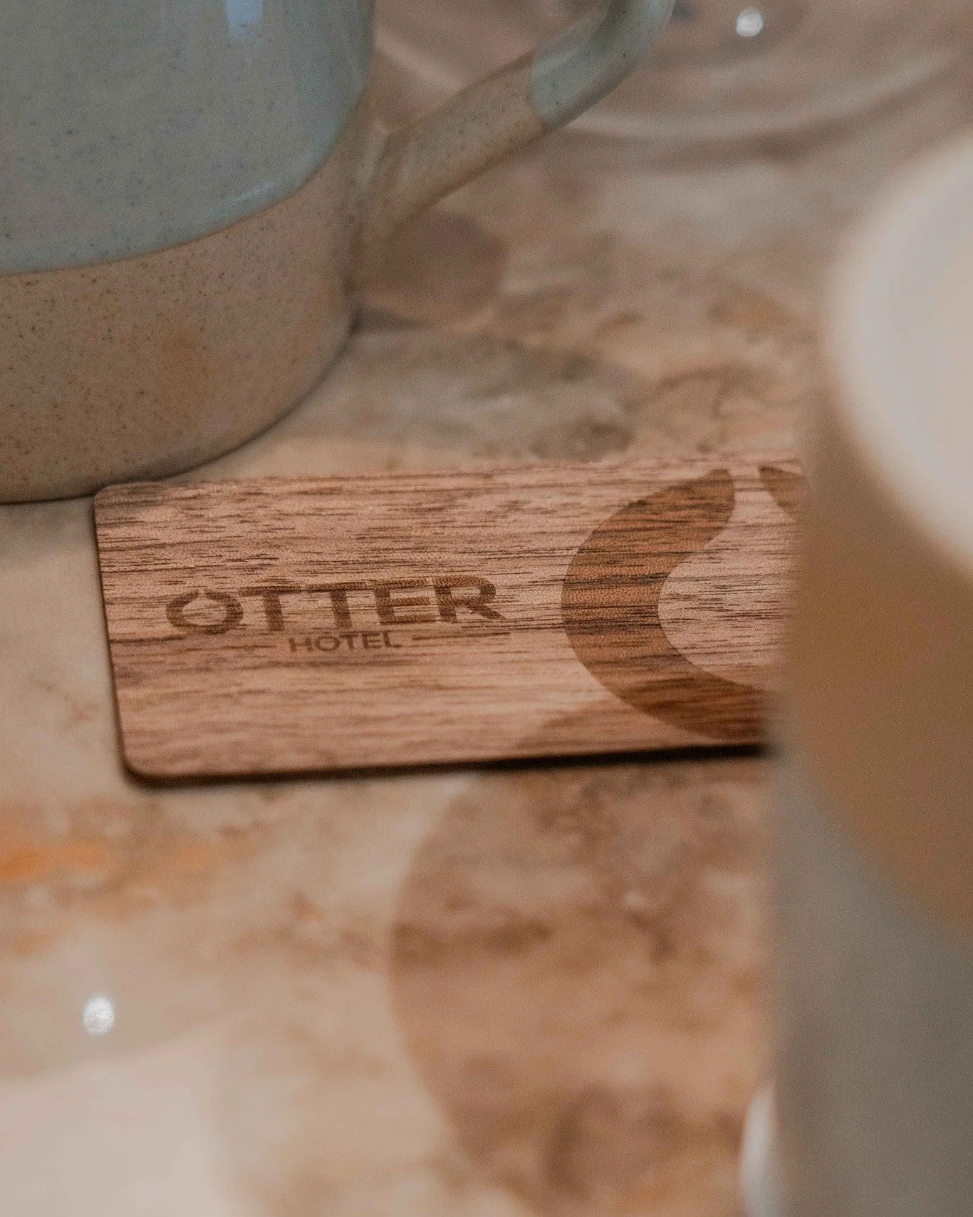 Wooden coaster engraved with the word “OTTER HOTEL” and an otter illustration on a marble tabletop.