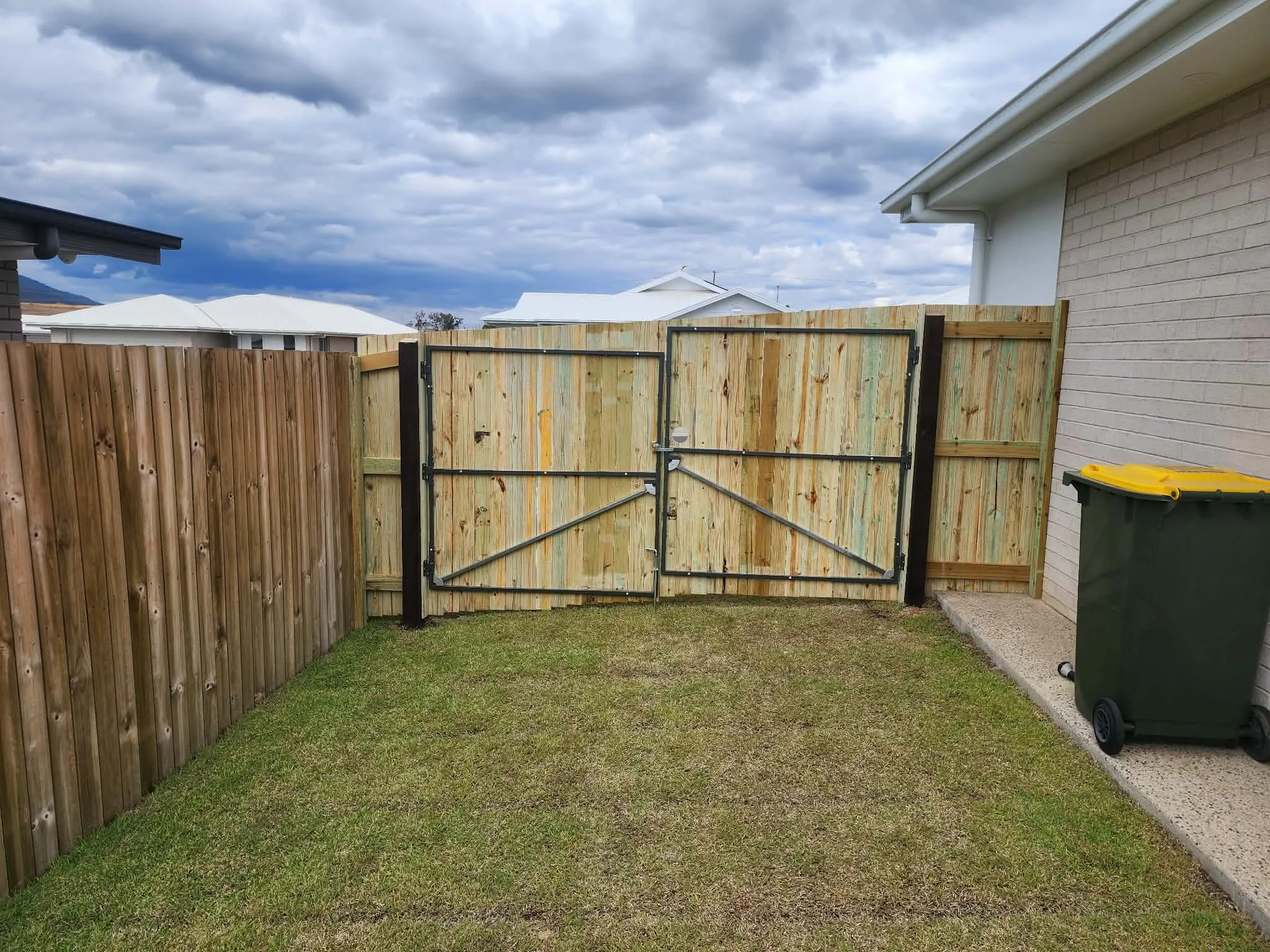 A backyard with a wooden privacy fence, green grass, and a partly cloudy sky, with neighboring houses visible in the background.