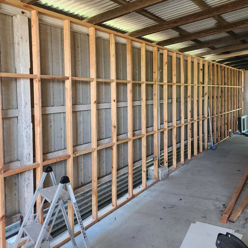 Unfinished interior wall with wooden framing and a corrugated metal ceiling, with construction tools including a ladder, a broom, and a small heater or vent.
