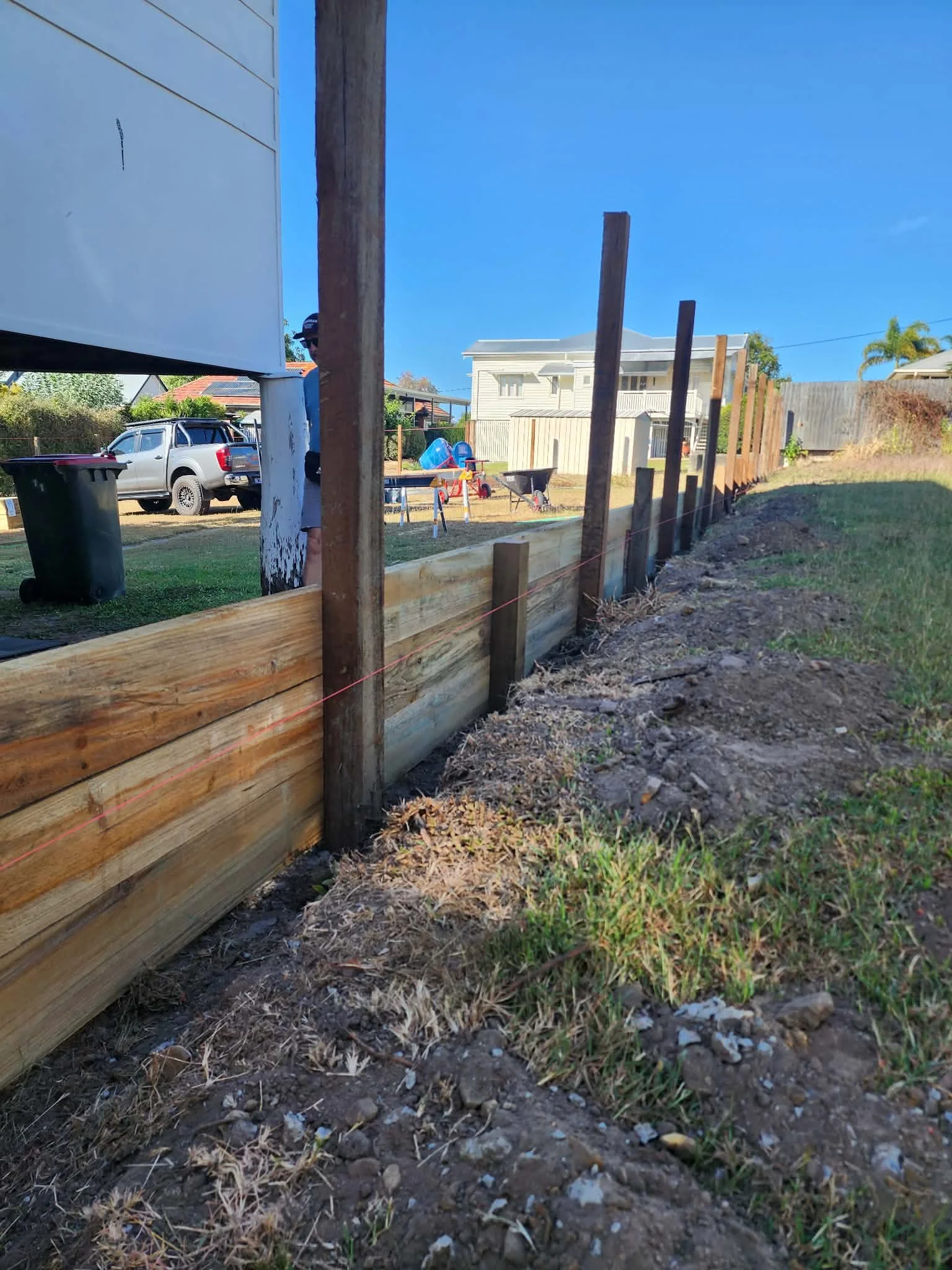 image shows a construction site with wooden fence posts and a horizontal wooden rail, a partially built fence in a backyard with houses, a vehicle, some tools, and trees in the background under a blue sky.