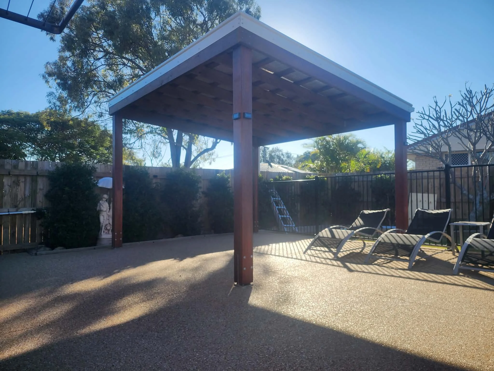 An outdoor backyard scene with a wooden deck structure, several lounge chairs, and a fence with trees and houses in the background.
