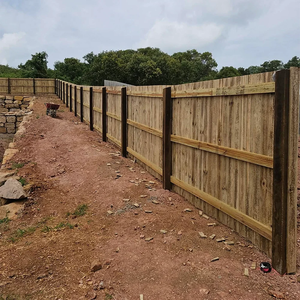 A newly built wooden fence along a dirt pathway with a wheelbarrow nearby, overgrown trees in the background, and a cloudy sky.