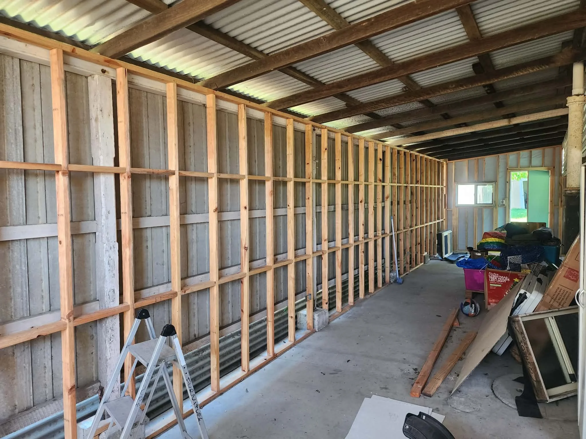 Interior of a house under renovation with exposed wooden framing and a corrugated metal roof. There's a small step ladder and various tools and supplies scattered on the floor.