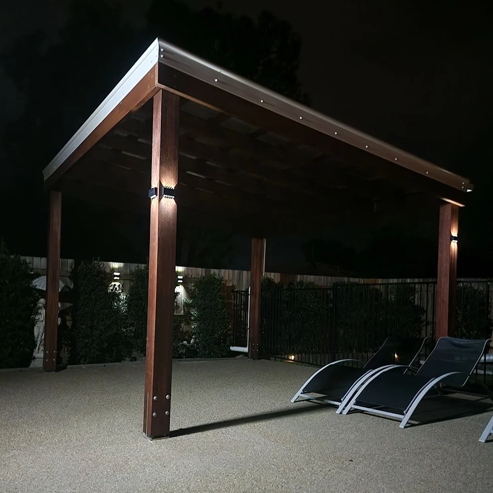 A covered outdoor patio with wooden support beams and a metal roof, with two lounge chairs on a textured concrete surface at night.