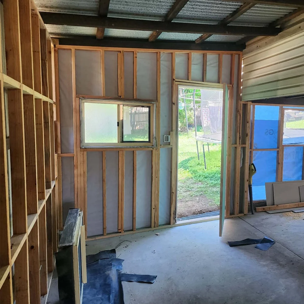 Interior of a house under construction with exposed wooden studs, a small window, and an open door leading outside to a grassy yard with a trampoline.