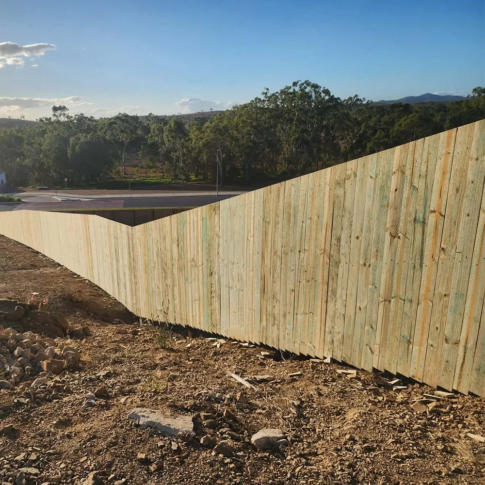 A long wooden fence with a sloped top runs alongside rocky dirt ground, with trees, a road, and hills in the background under a partly cloudy sky.