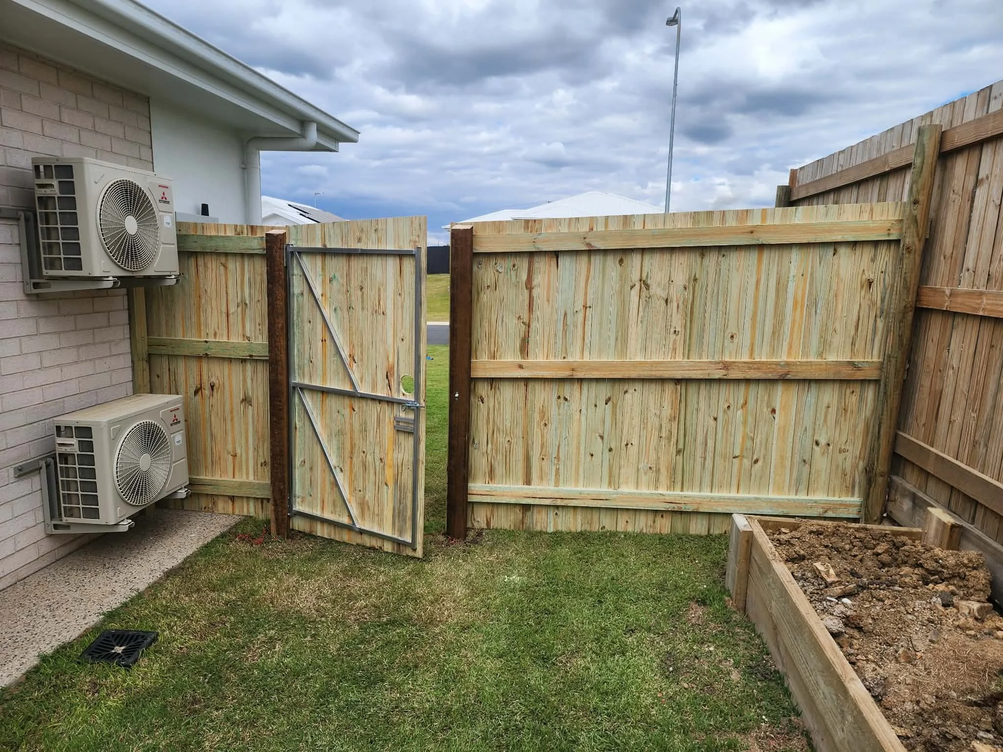 A backyard enclosed by a tall wooden fence. The sky is partly cloudy. On the left side, there are two air conditioning units mounted on the house wall. A gate in the fence is open, leading to a grassy area and neighboring rooftops beyond. In the bottom right corner, there is a planter box filled with soil, and in the bottom left corner, a drain on the grass.