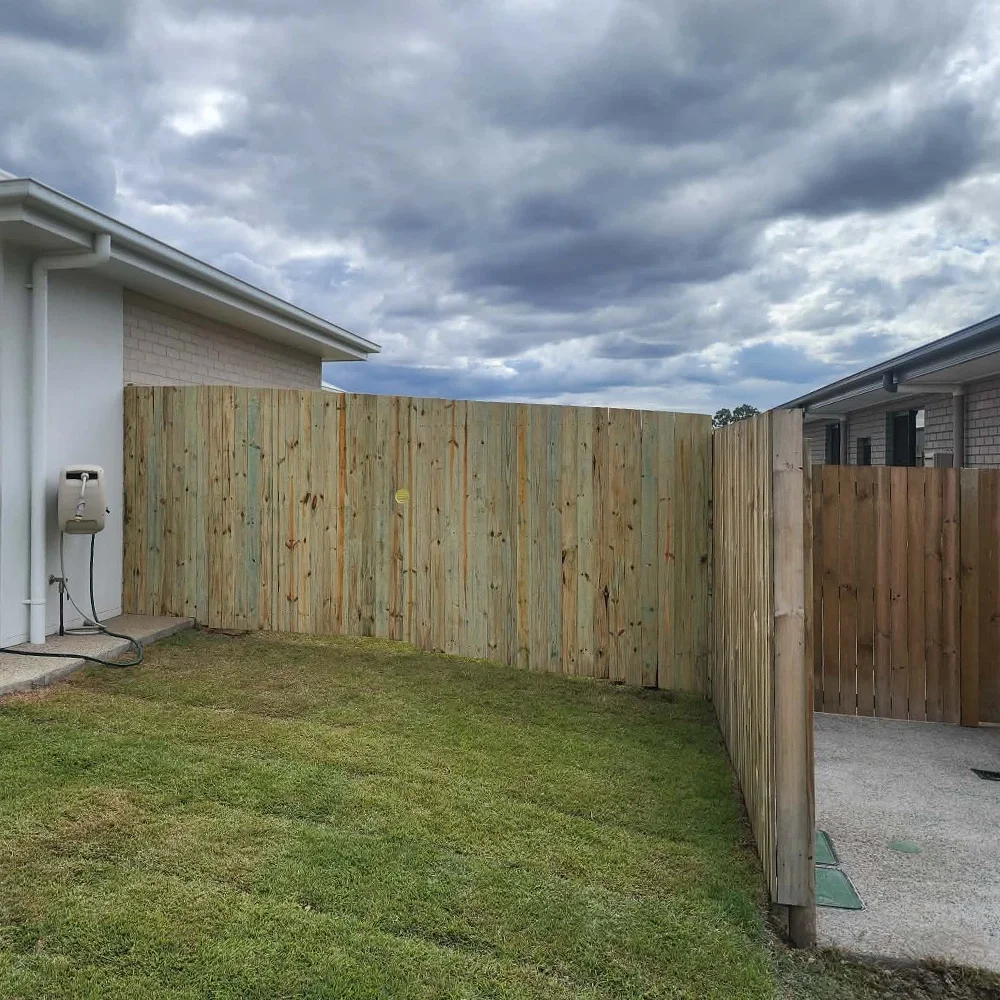 A backyard with a new wooden privacy fence, part of a house with a white exterior, and a cloudy sky overhead.
