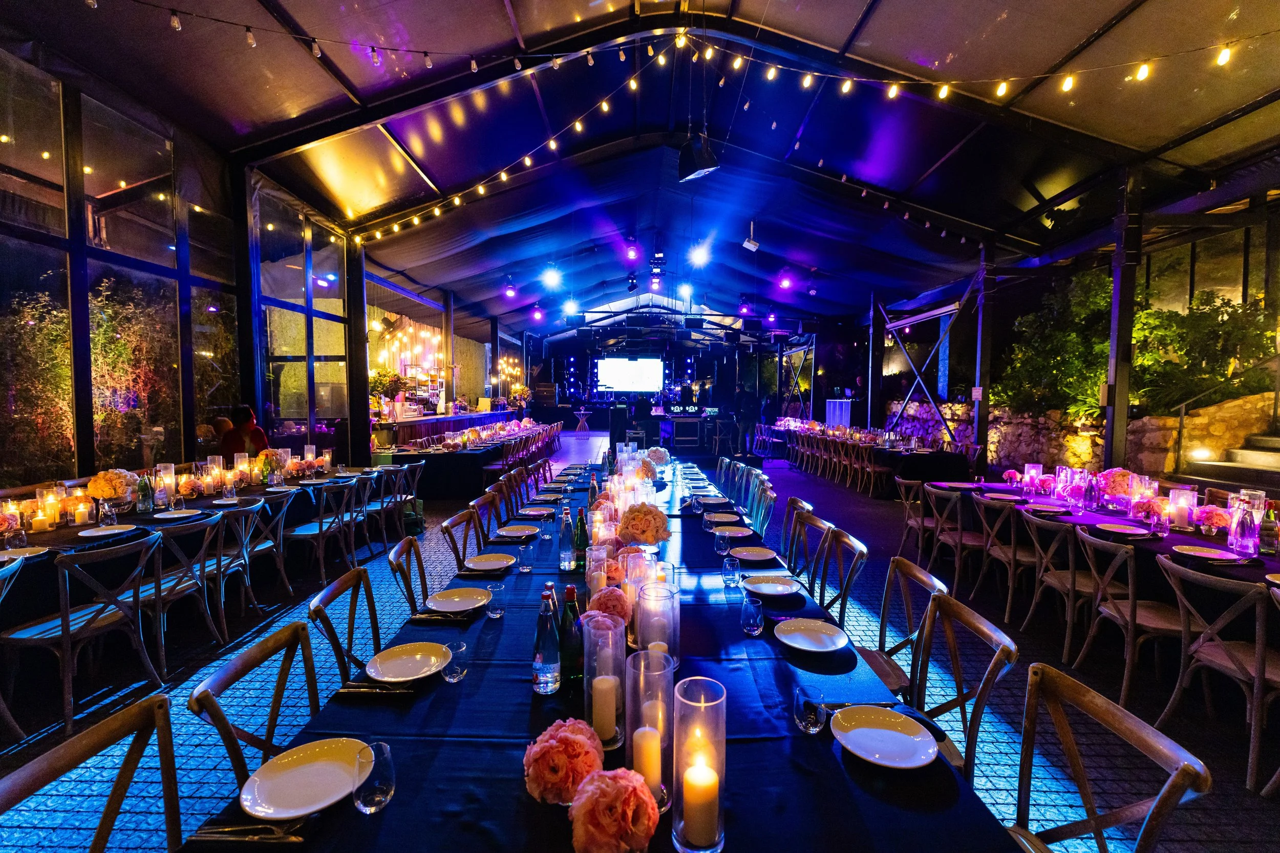 Wedding reception with long tables decorated with pink flowers and candles, under a tent with string lights and purple stage lighting, set outdoors at night.