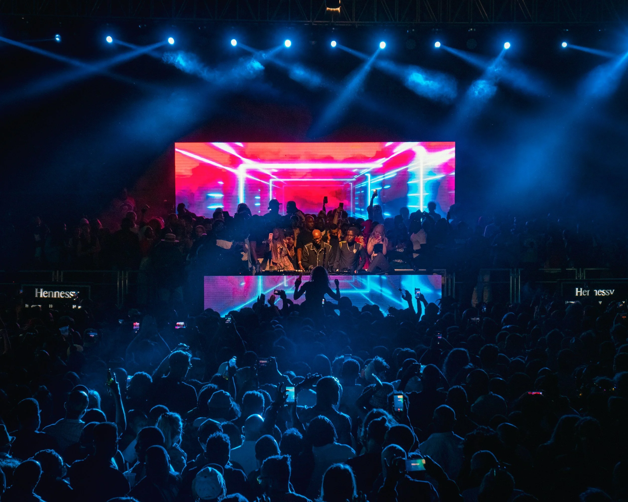 A lively concert scene with a large audience watching a DJ perform on stage. The stage features colorful LED screens displaying abstract visuals with neon lights and smoke effects. The crowd is energetic, with some people raising their hands and recording the performance on their phones.